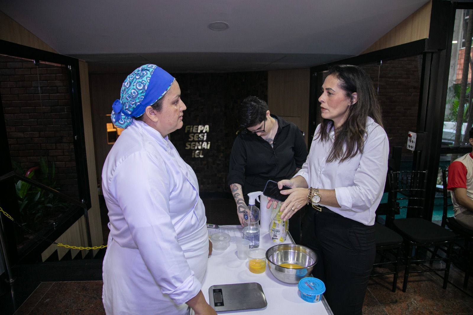 Três pessoas em uma cozinha. Uma chef com lenço na cabeça, uma chef e uma mulher sentadas à mesa com ingredientes, possivelmente conversando.