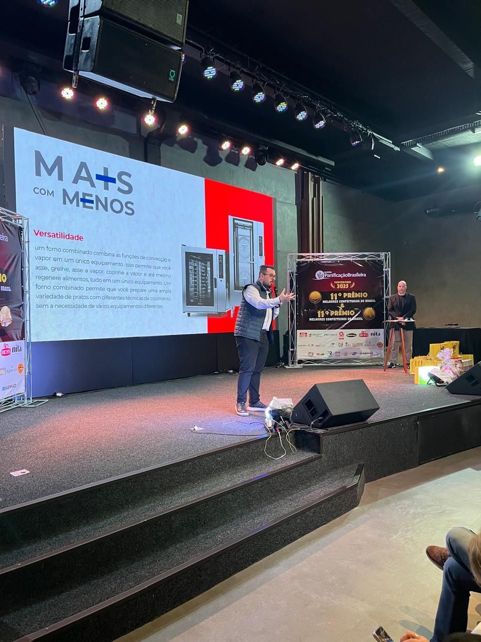 Homem apresentando em um palco com uma tela exibindo o texto 