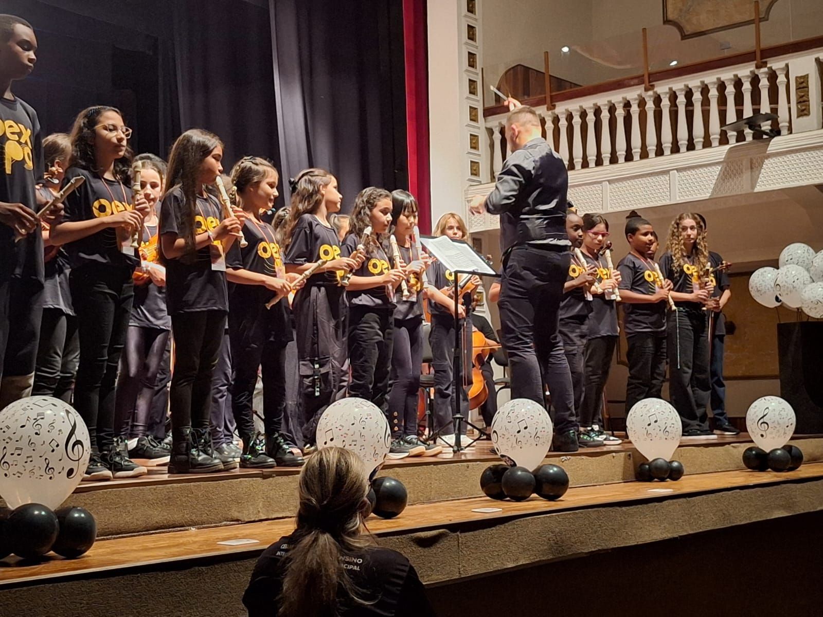 Um grupo musical infantil se apresenta no palco sob a regência de um maestro, e balões decoram o palco.