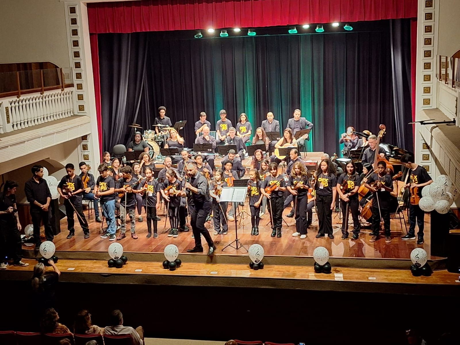 Uma orquestra se apresenta no palco, com os músicos segurando seus instrumentos. O palco está escuro, com luzes suspensas.