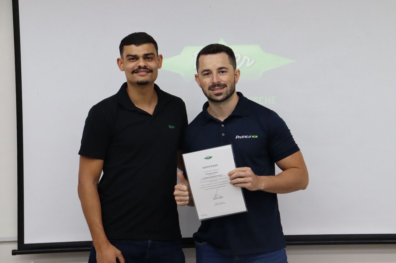 Dois homens em pé, sorrindo, um deles segurando um papel; fundo com logotipo.