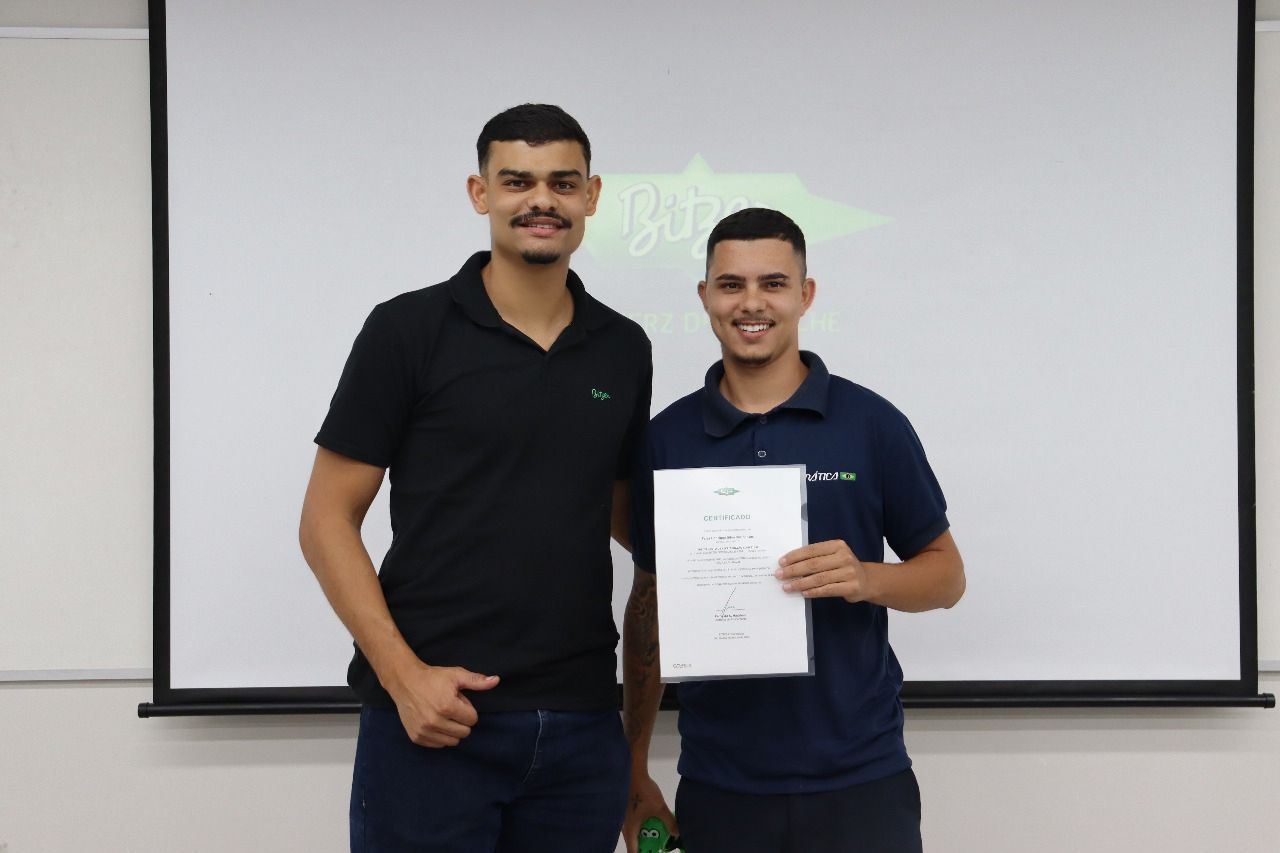 Dois homens sorrindo, um deles segurando um certificado em frente a uma tela de projeção em uma sala.