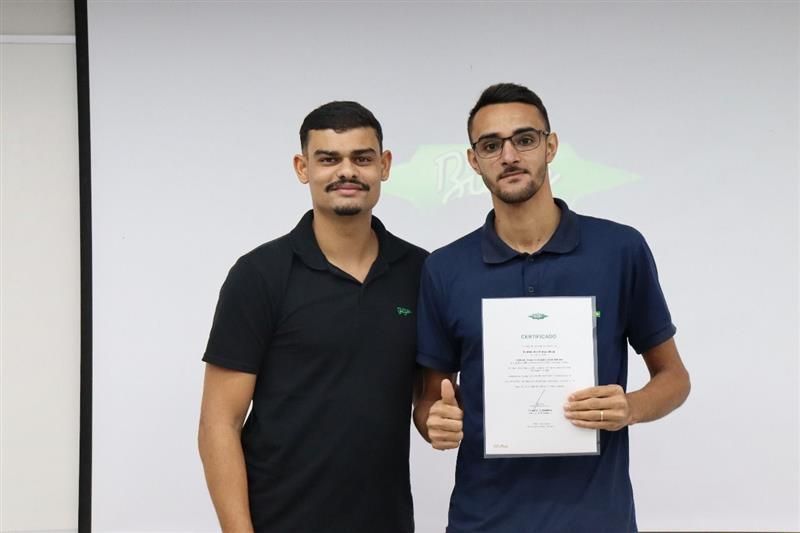 Dois homens em frente a uma tela, um deles segurando um certificado, fazendo um sinal de positivo com o polegar.