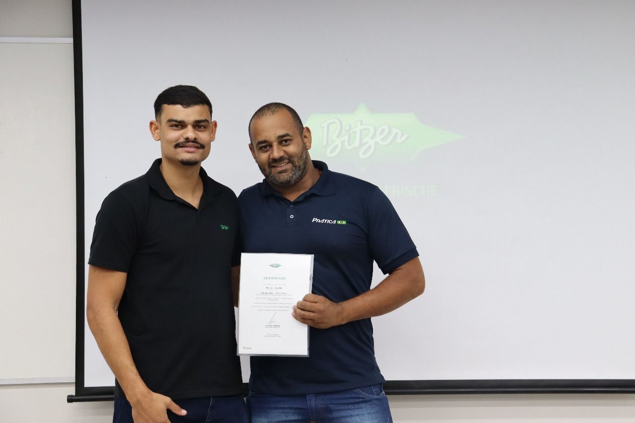 Dois homens estão de pé, um deles segurando um certificado em frente a uma tela de projeção; ambos sorriem.