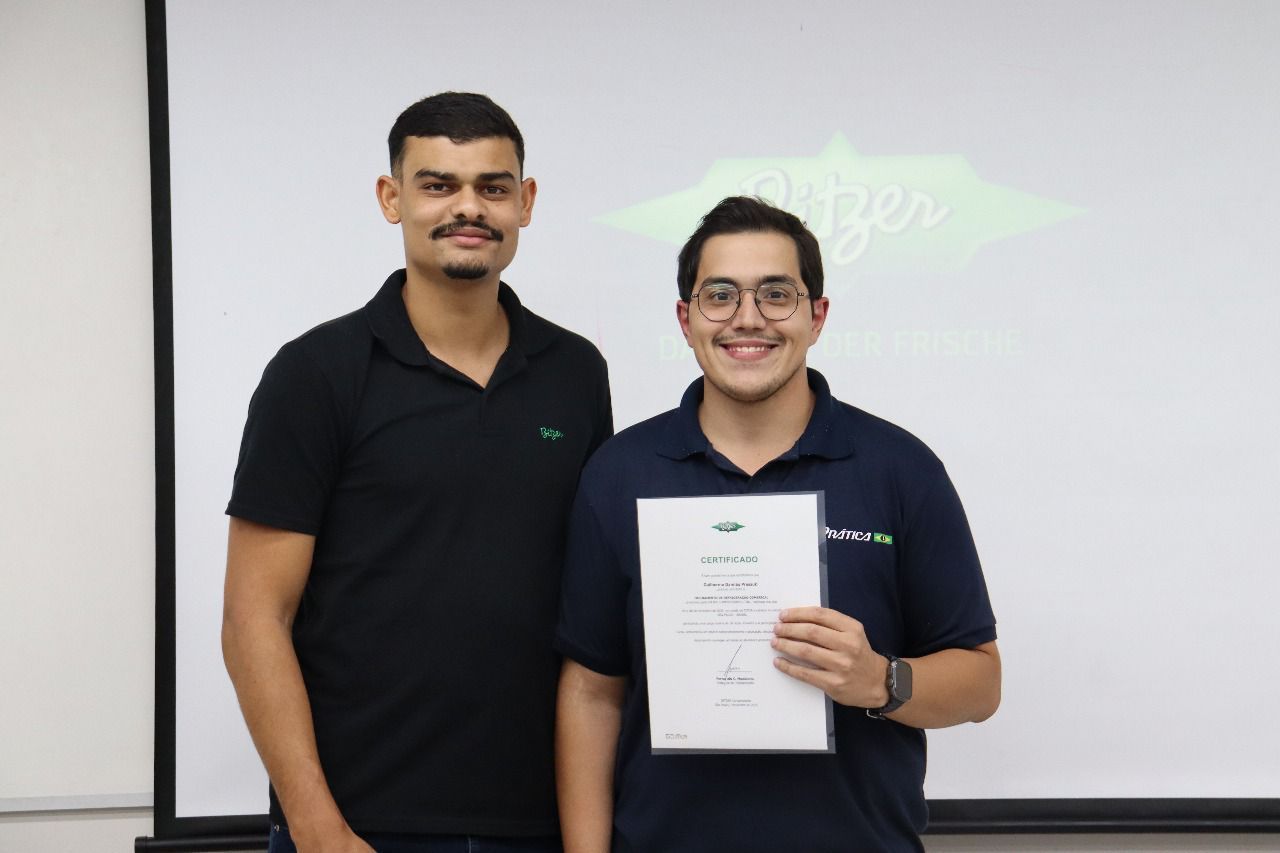 Dois homens estão em frente a uma tela. Um deles segura um certificado, e ambos sorriem. O local parece ser um escritório.