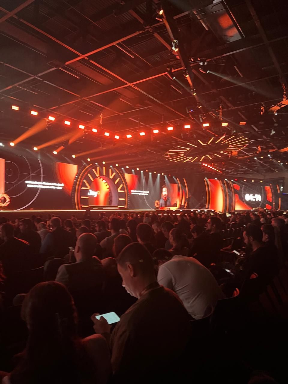 Grande auditório, palco iluminado com luzes vermelhas, público sentado, evento em andamento.