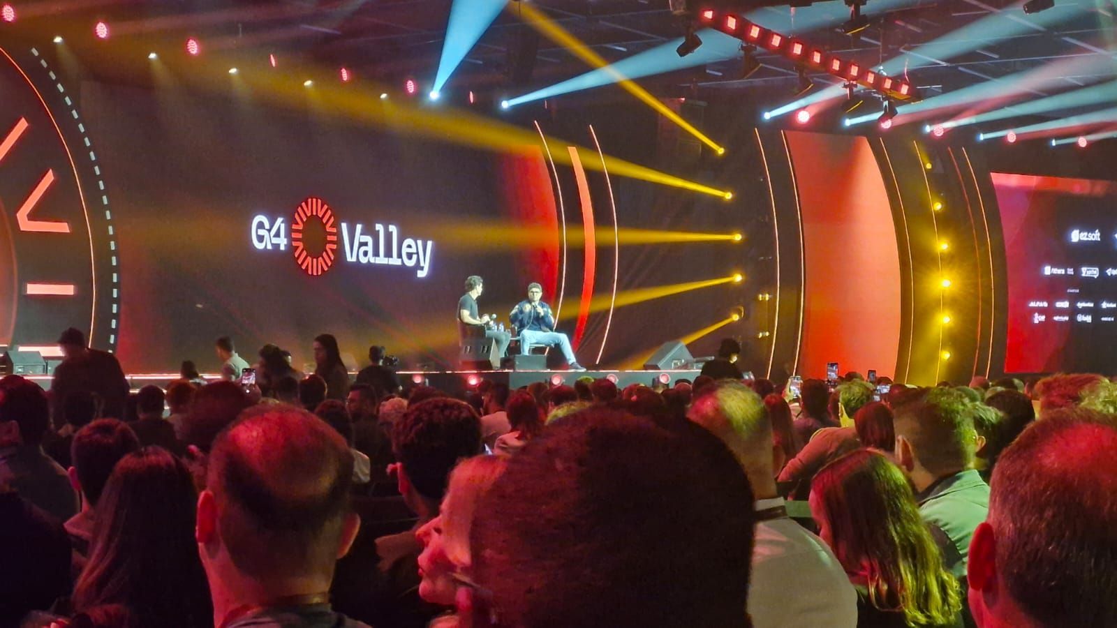 O público em uma conferência observa duas pessoas no palco, iluminadas por luzes de palco; o logotipo