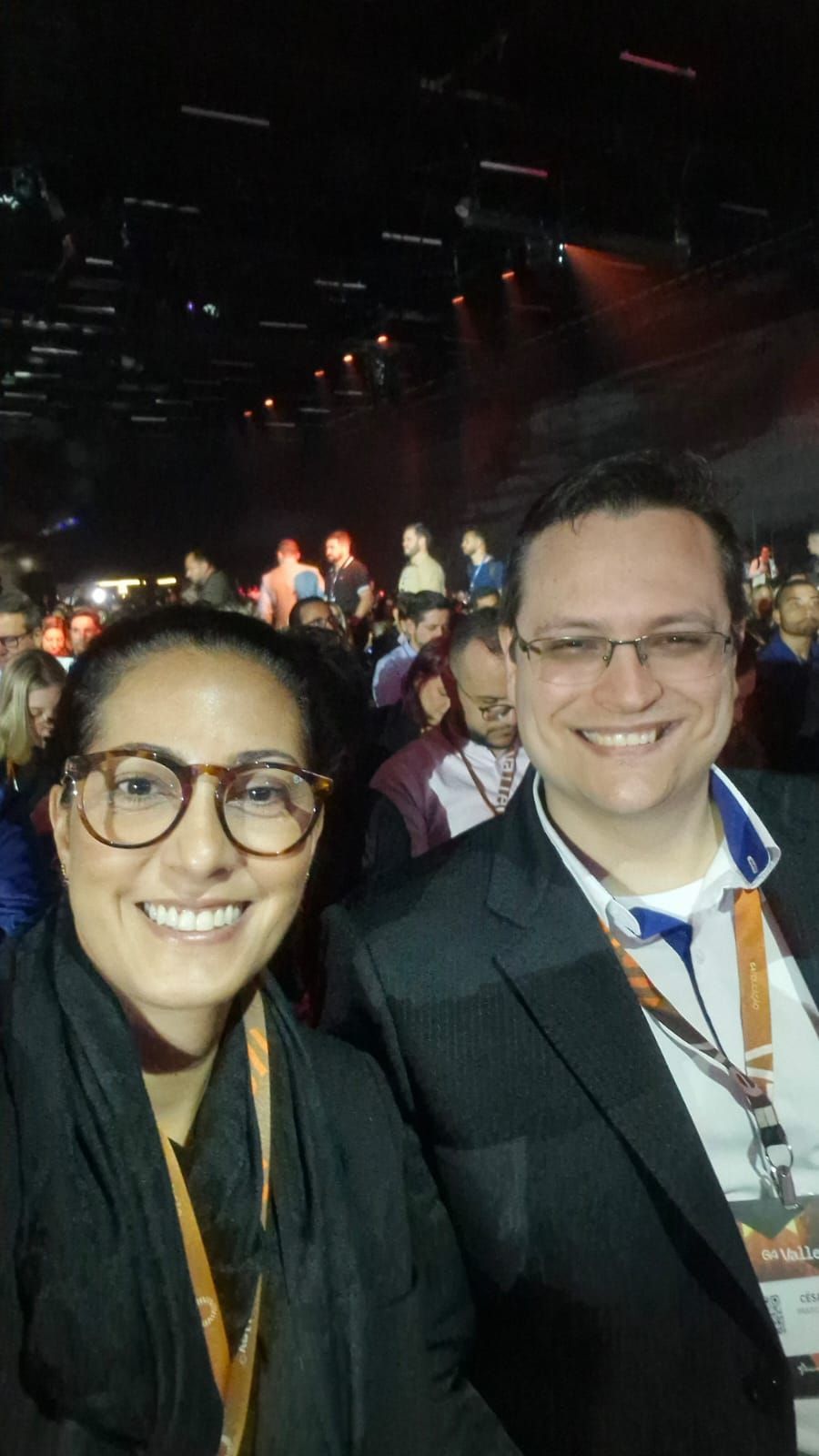 Mulher e homem sorrindo para a câmera, pessoas ao fundo, ambiente iluminado.