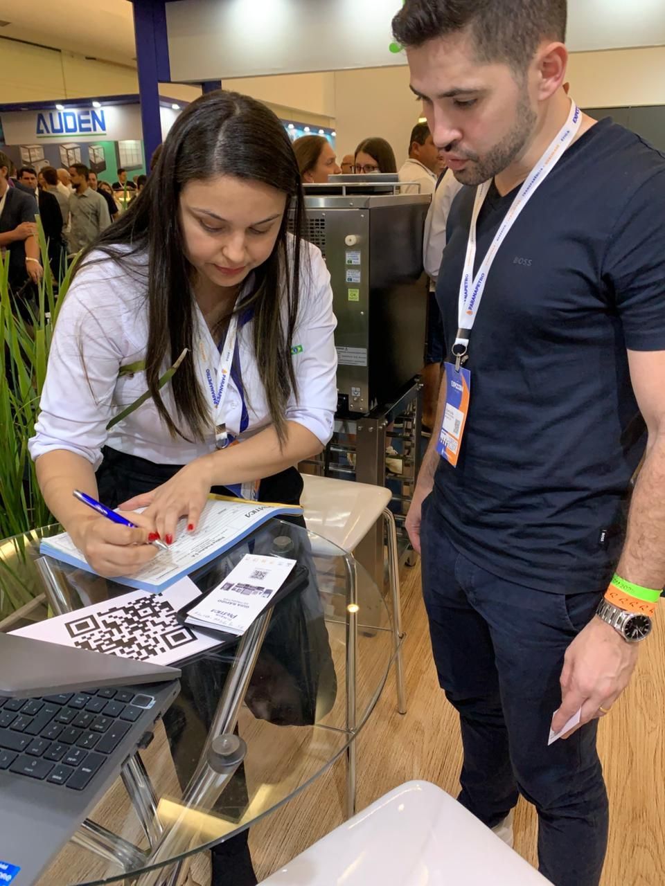 Mulher assinando um formulário enquanto um homem observa. Eles estão em uma cabine com um computador e códigos QR.