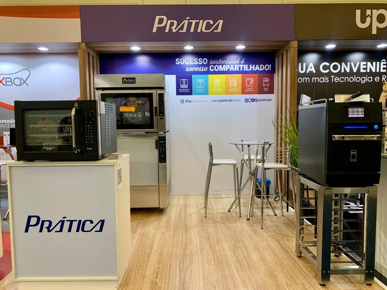 Exposição de equipamentos de cozinha da Prática em uma feira comercial. Apresenta fornos e uma máquina de café em um ambiente bem iluminado.