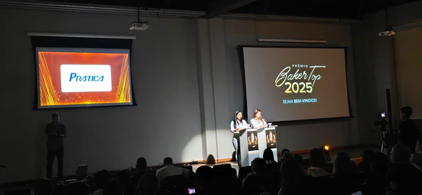 Duas mulheres no palco apresentam em uma conferência. Duas telas exibem logotipos e texto, com uma plateia em primeiro plano.