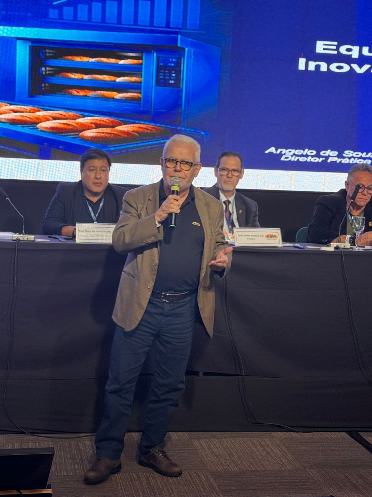 Homem discursando em uma conferência. Outras pessoas sentadas à mesa atrás dele; uma tela exibe produtos de confeitaria.