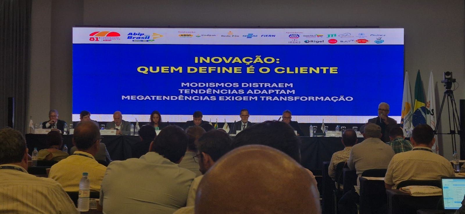 Apresentação em conferência sobre inovação. Um fundo azul exibe o texto. Público sentado, palestrantes em uma mesa.