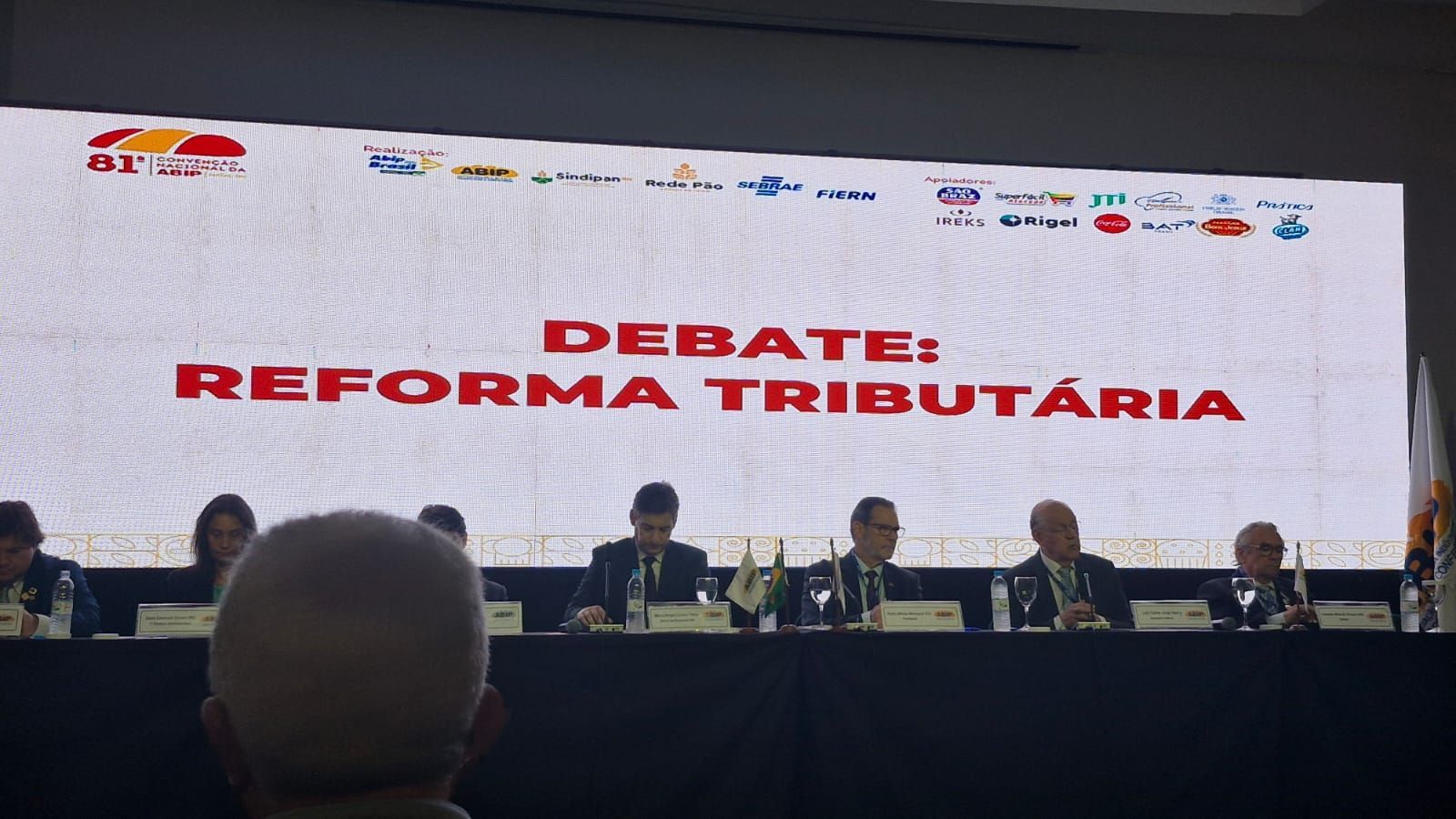 Debate sobre reforma tributária. Palestrantes sentados à mesa, tela exibe 