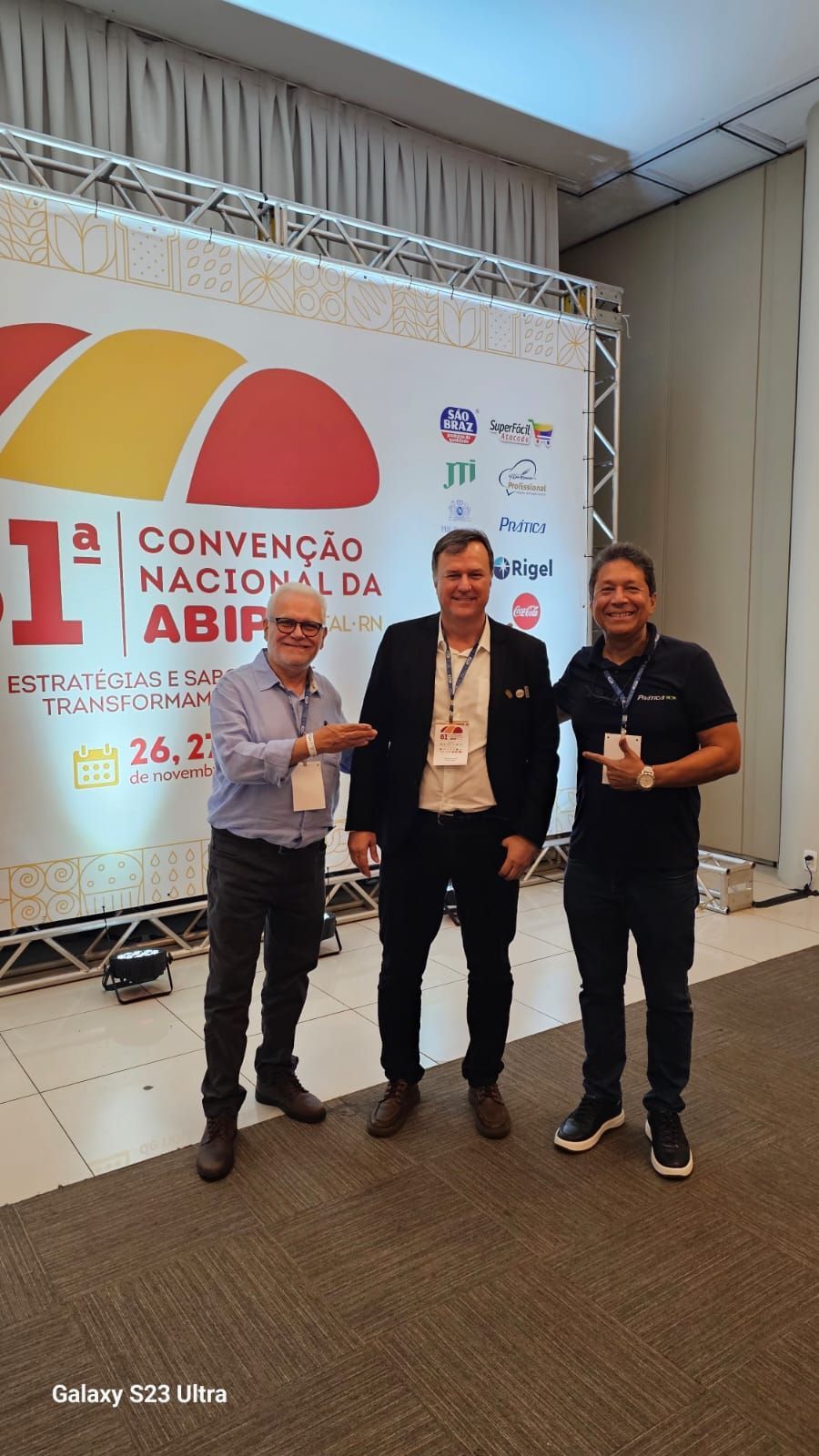 Três homens estão em pé em frente a uma faixa de uma conferência. O homem à esquerda aponta. Eles sorriem em um ambiente de convenção.
