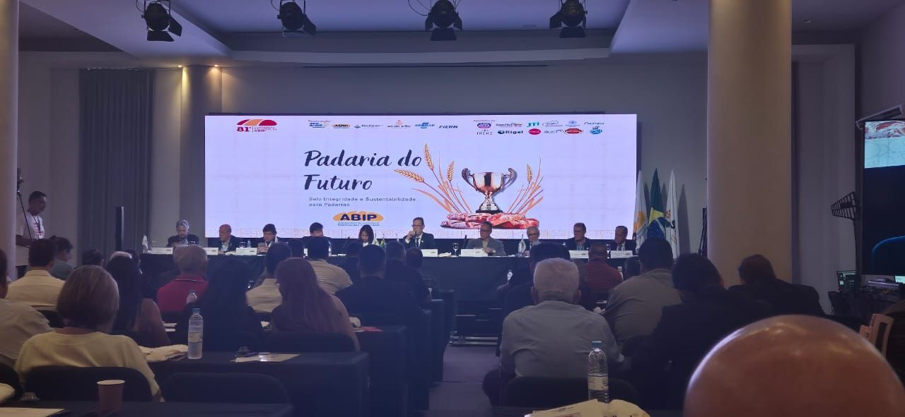 Público em um auditório assistindo a uma apresentação no palco, com um telão exibindo o título.