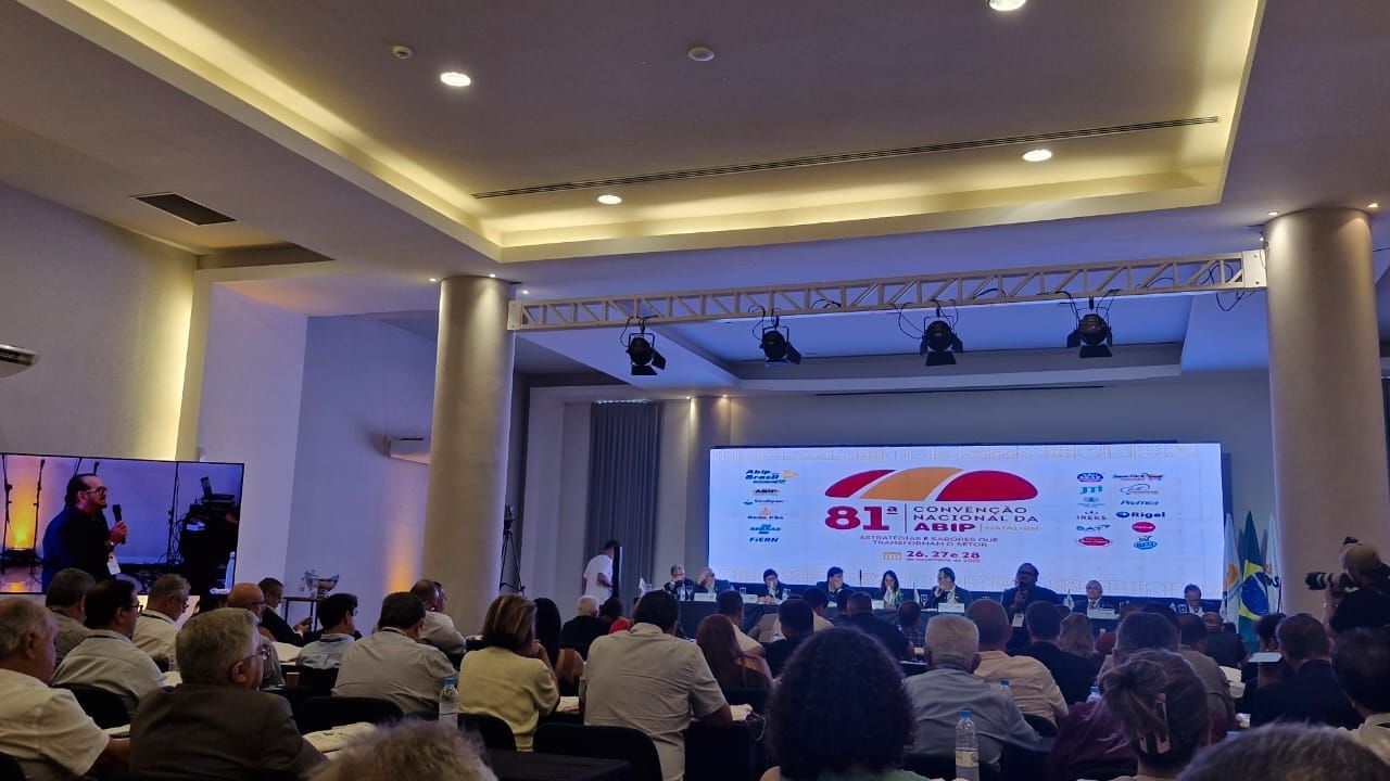 Conferência em um grande salão com um palestrante no pódio e o público sentado em mesas. Um telão exibe informações sobre o evento.