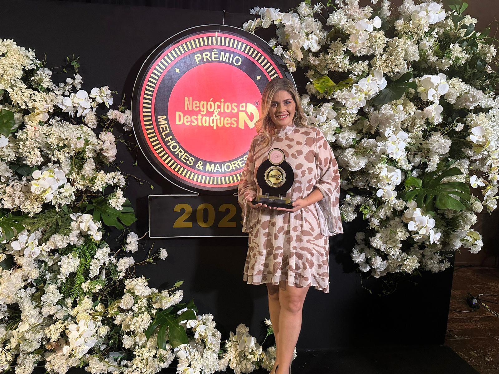 Mulher de vestido, segurando um prêmio, posa em frente a uma placa e um arranjo floral.