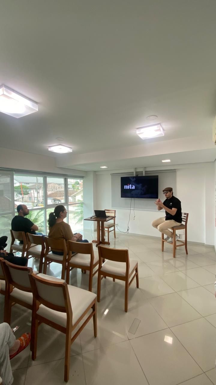 Homem fazendo uma apresentação para uma plateia sentada em uma sala bem iluminada. A tela exibe um logotipo; cadeiras dispostas em fila.