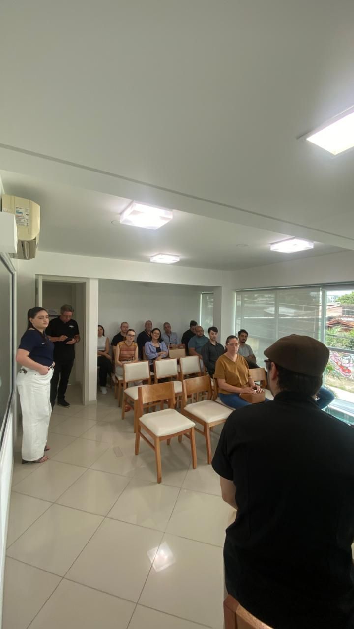 Pessoas em uma reunião, algumas sentadas em cadeiras, outras em pé. Paredes brancas, luzes fortes no teto, janela de vidro.