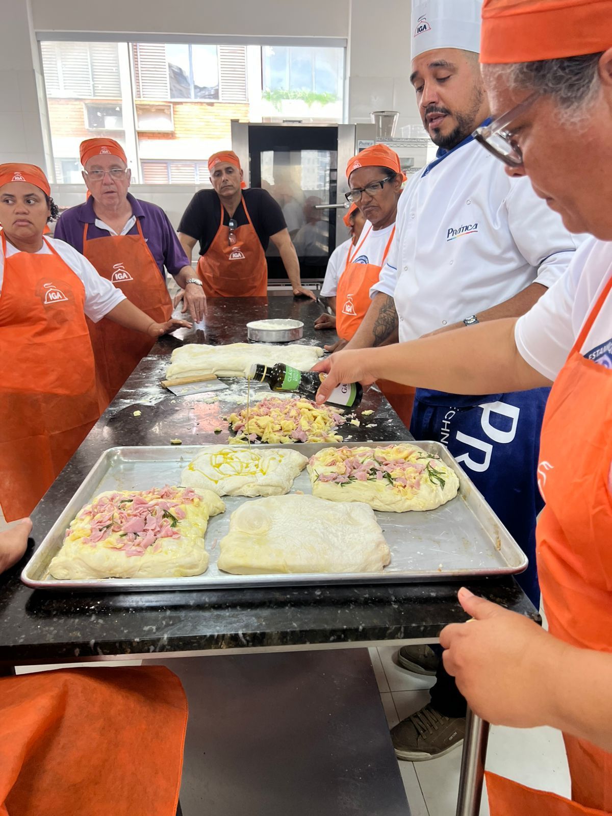 Aula de culinária: o chef demonstra como montar um pão, enquanto os alunos observam. Forno, mesa preta, aventais laranja.