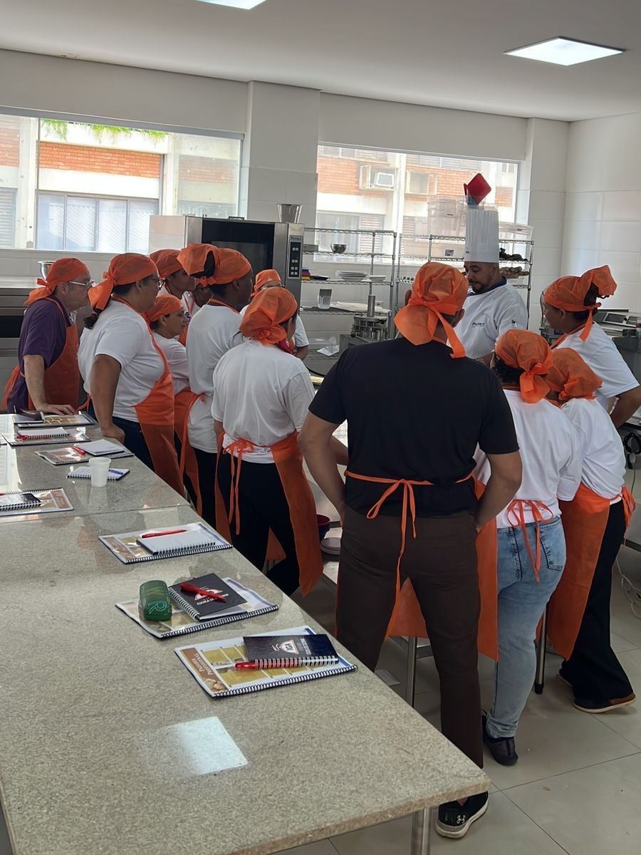 Uma aula de culinária em uma cozinha. Alunos e chef usam aventais e lenços na cabeça, reunidos em volta de uma bancada.