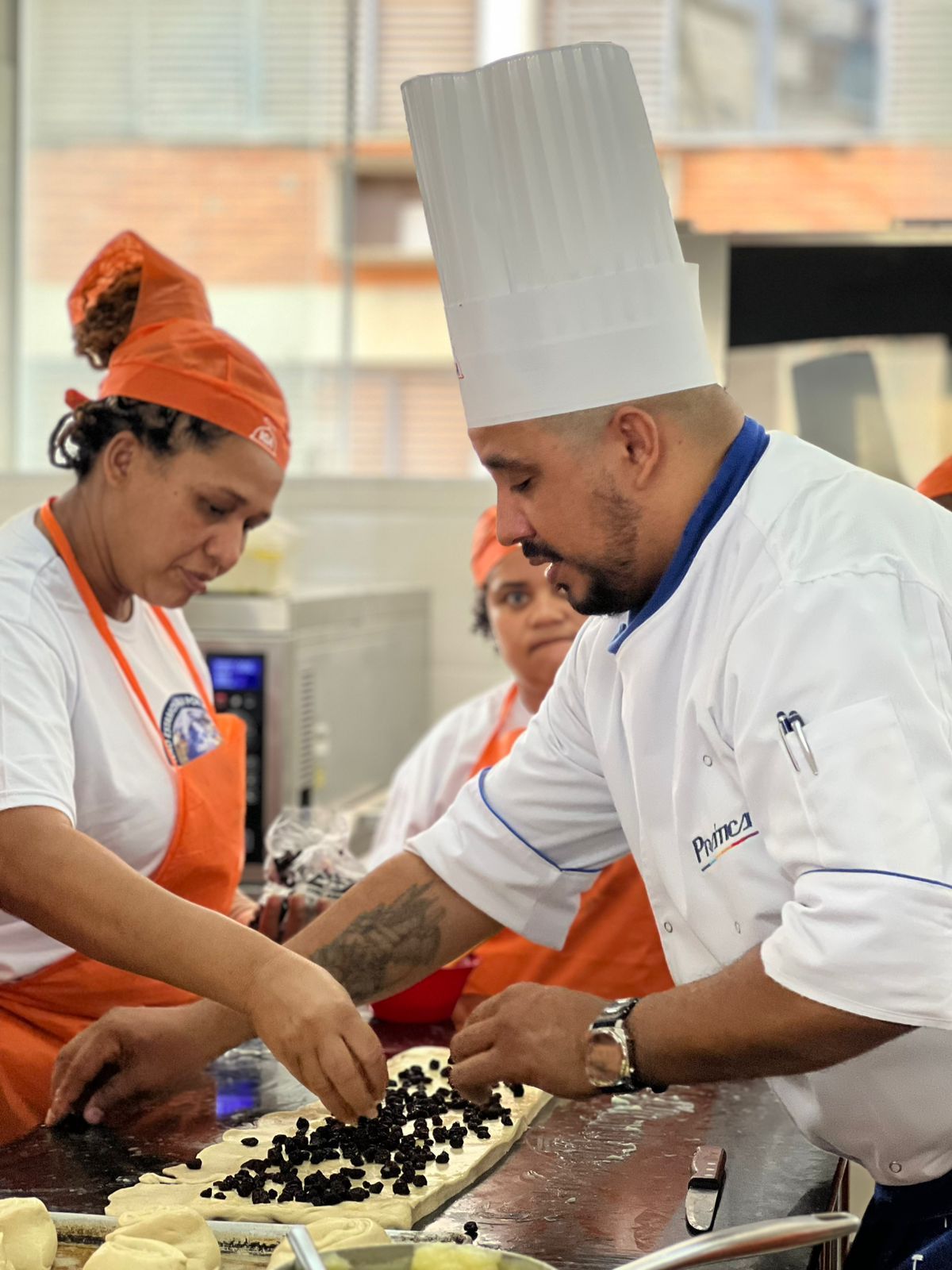 O chef adiciona ingredientes à massa com outras duas pessoas na cozinha.