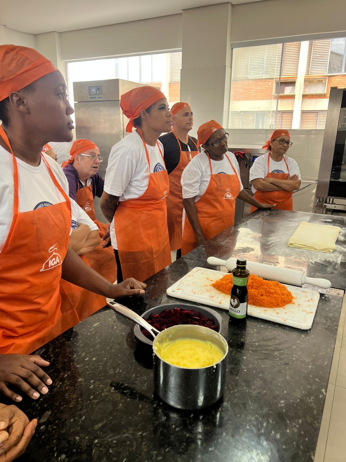 Grupo de pessoas com aventais e lenços na cabeça cor de laranja, assistindo a uma demonstração culinária em uma cozinha.