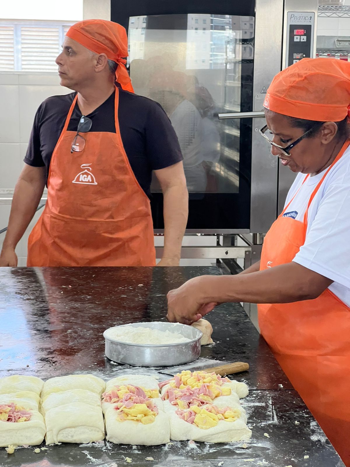 Duas pessoas com aventais e toucas laranja preparando comida em uma cozinha.