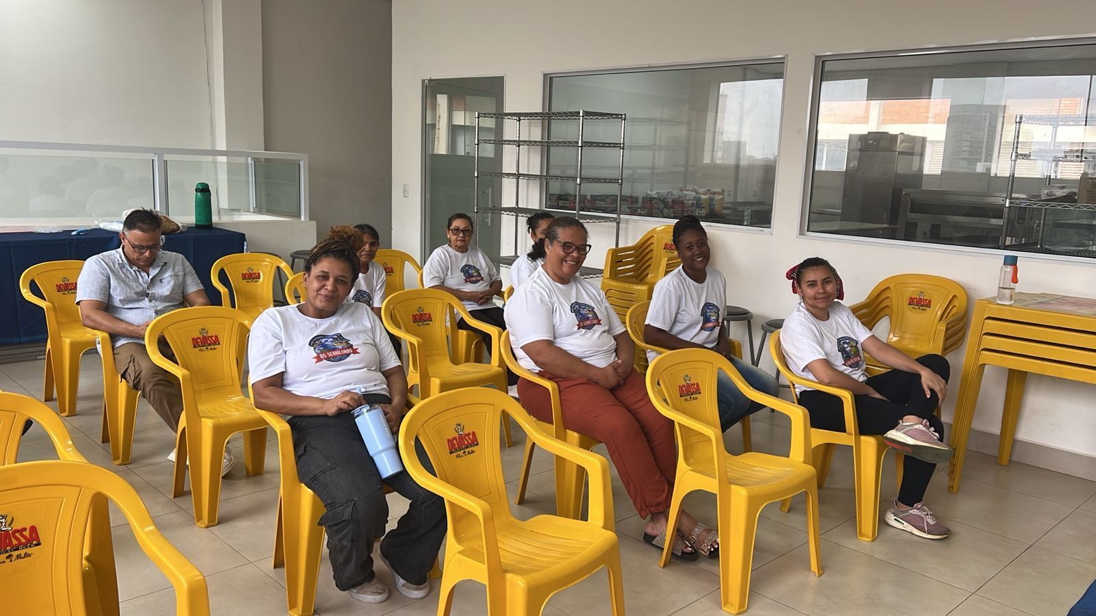 Grupo de pessoas sentadas em cadeiras amarelas, algumas vestindo camisas iguais, em uma sala com janelas.