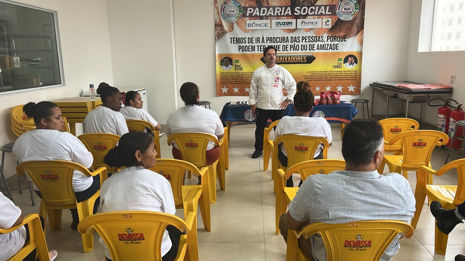Pessoas sentadas, ouvindo um homem falar em uma apresentação, faixa na parede: