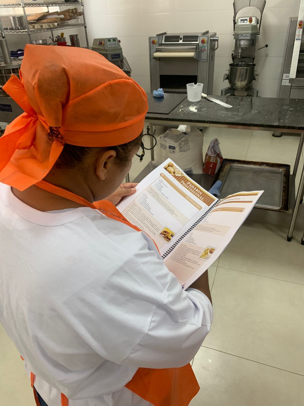 Pessoa com lenço laranja na cabeça e avental lendo uma receita em uma cozinha comercial.