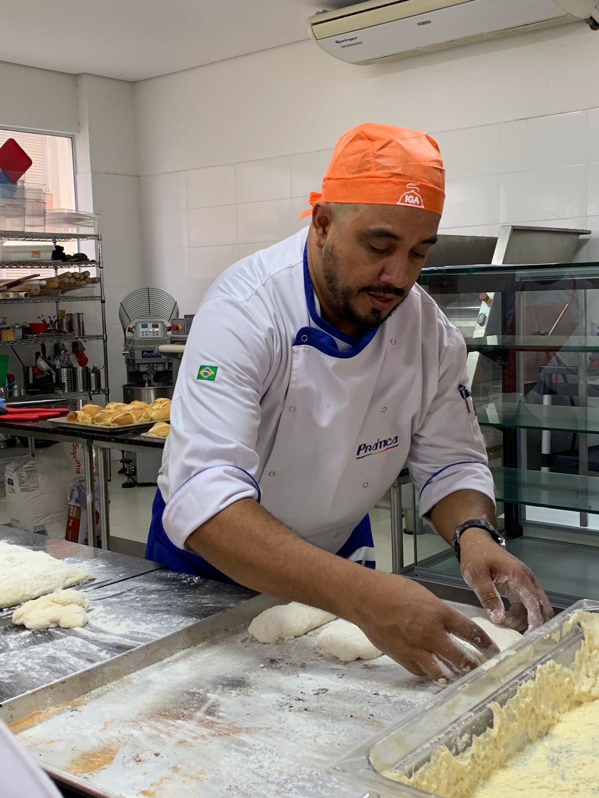 Chef moldando a massa em uma cozinha; ele veste um jaleco branco e um chapéu laranja.