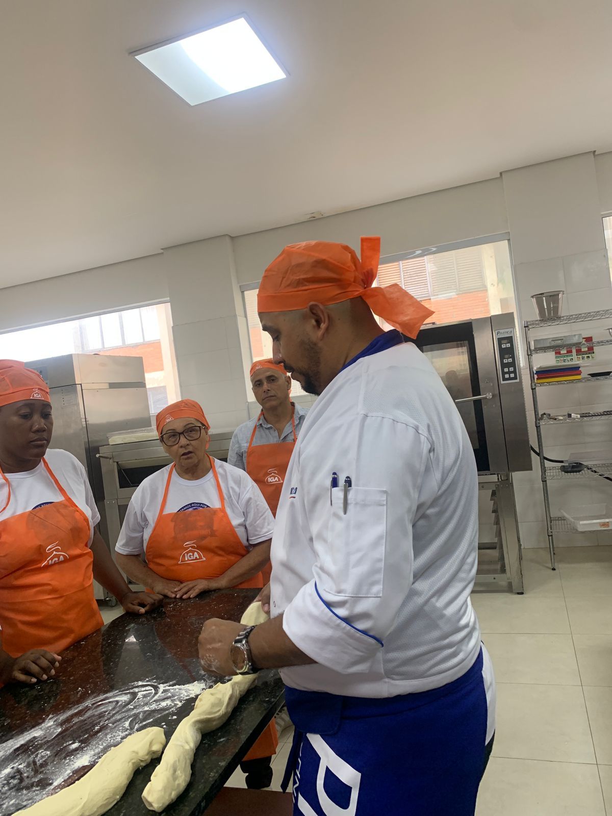 Padeiro de jaleco branco moldando a massa enquanto outros, de uniforme laranja, observam em uma padaria.