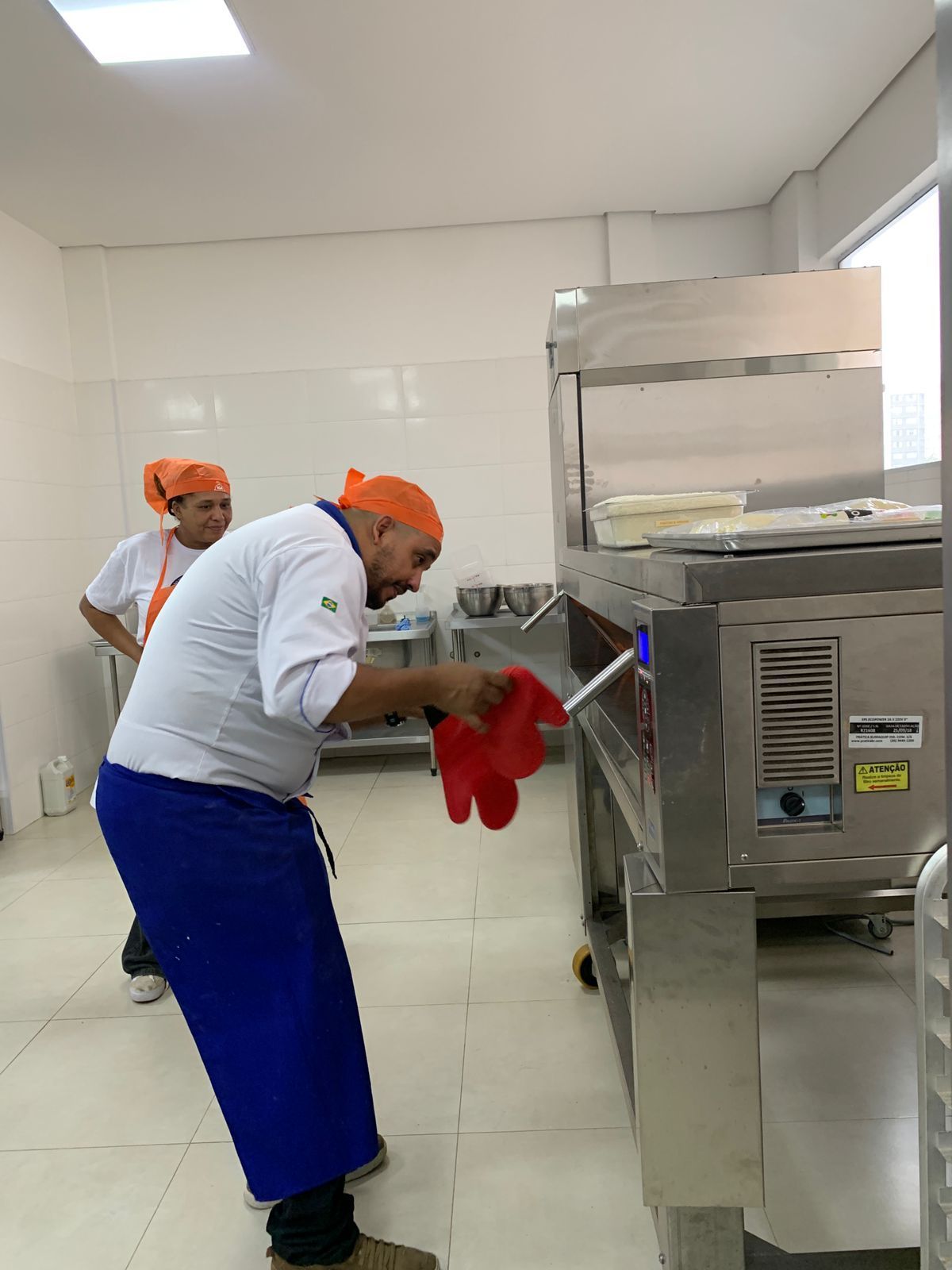 Um chef de cozinha usa luvas de forno vermelhas para retirar comida do forno. Outra pessoa observa.