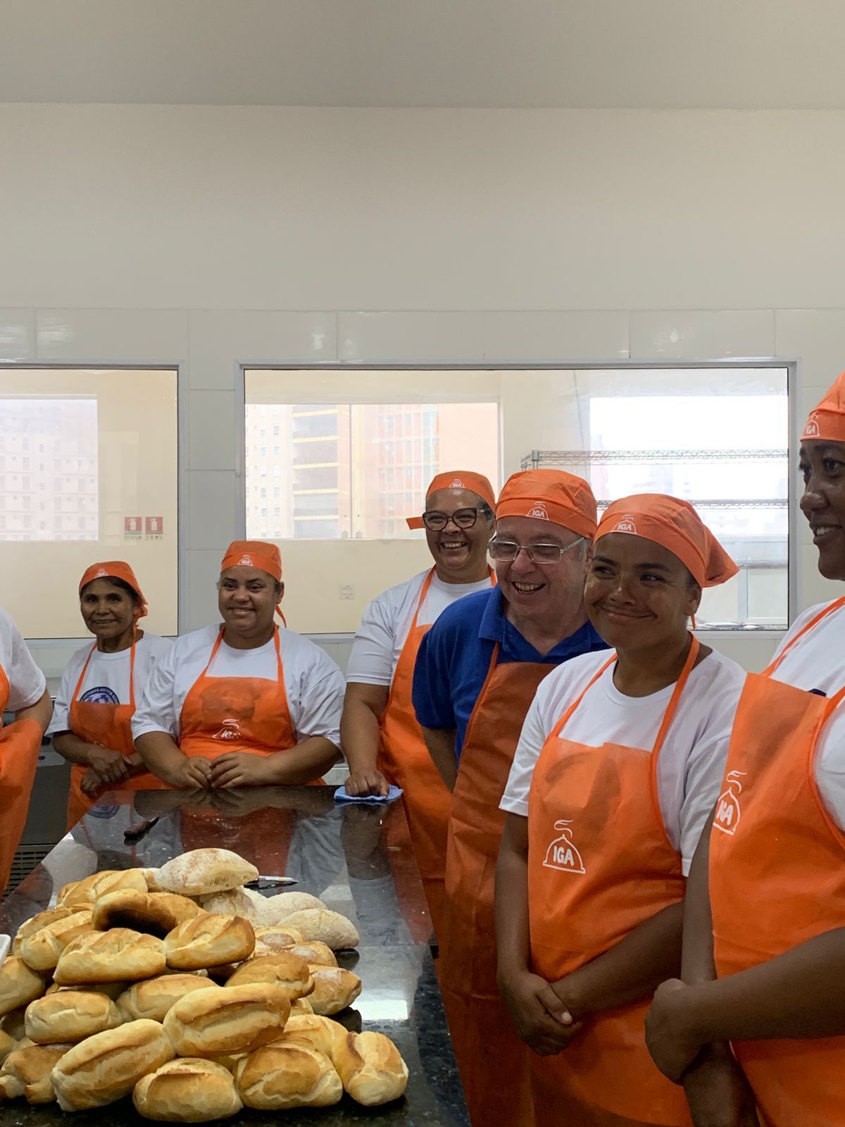 Pessoas usando aventais e lenços na cabeça na cor laranja, sorrindo perto de pães assados em um balcão.