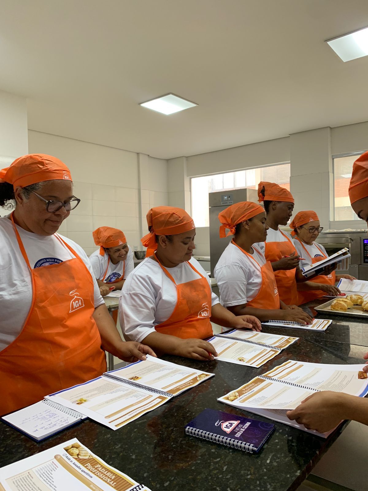 Pessoas com aventais e lenços na cabeça, na cor laranja, em uma cozinha, olhando documentos.