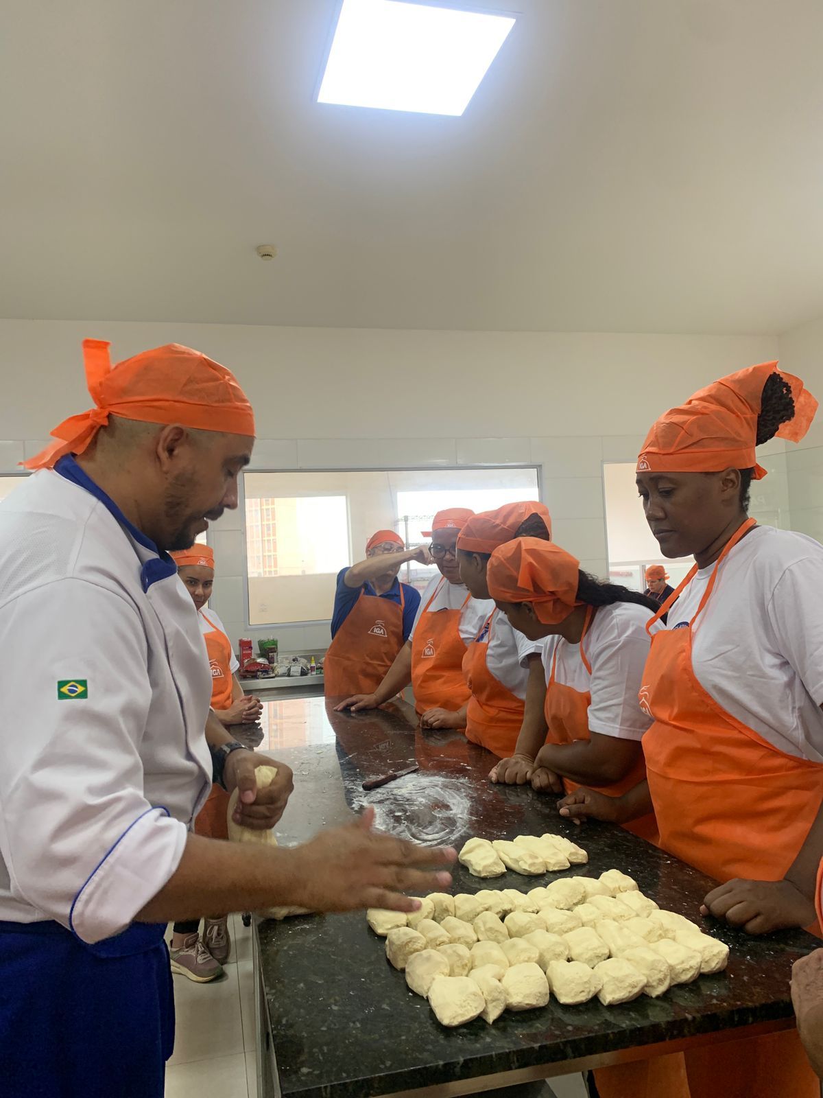 Um chef orienta um grupo na preparação de massa em uma cozinha. Eles usam aventais e lenços na cabeça na cor laranja.