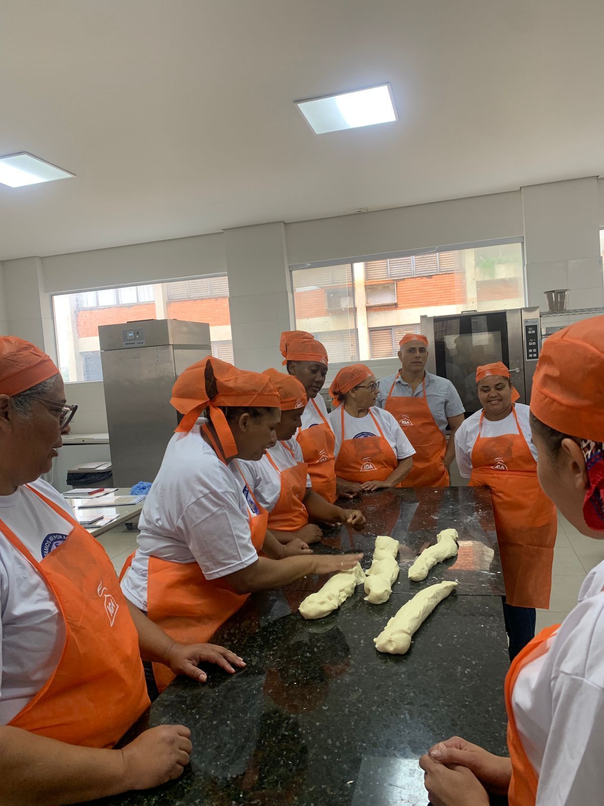 Grupo de pessoas com aventais e lenços na cabeça cor de laranja, abrindo massa em uma bancada escura na cozinha.
