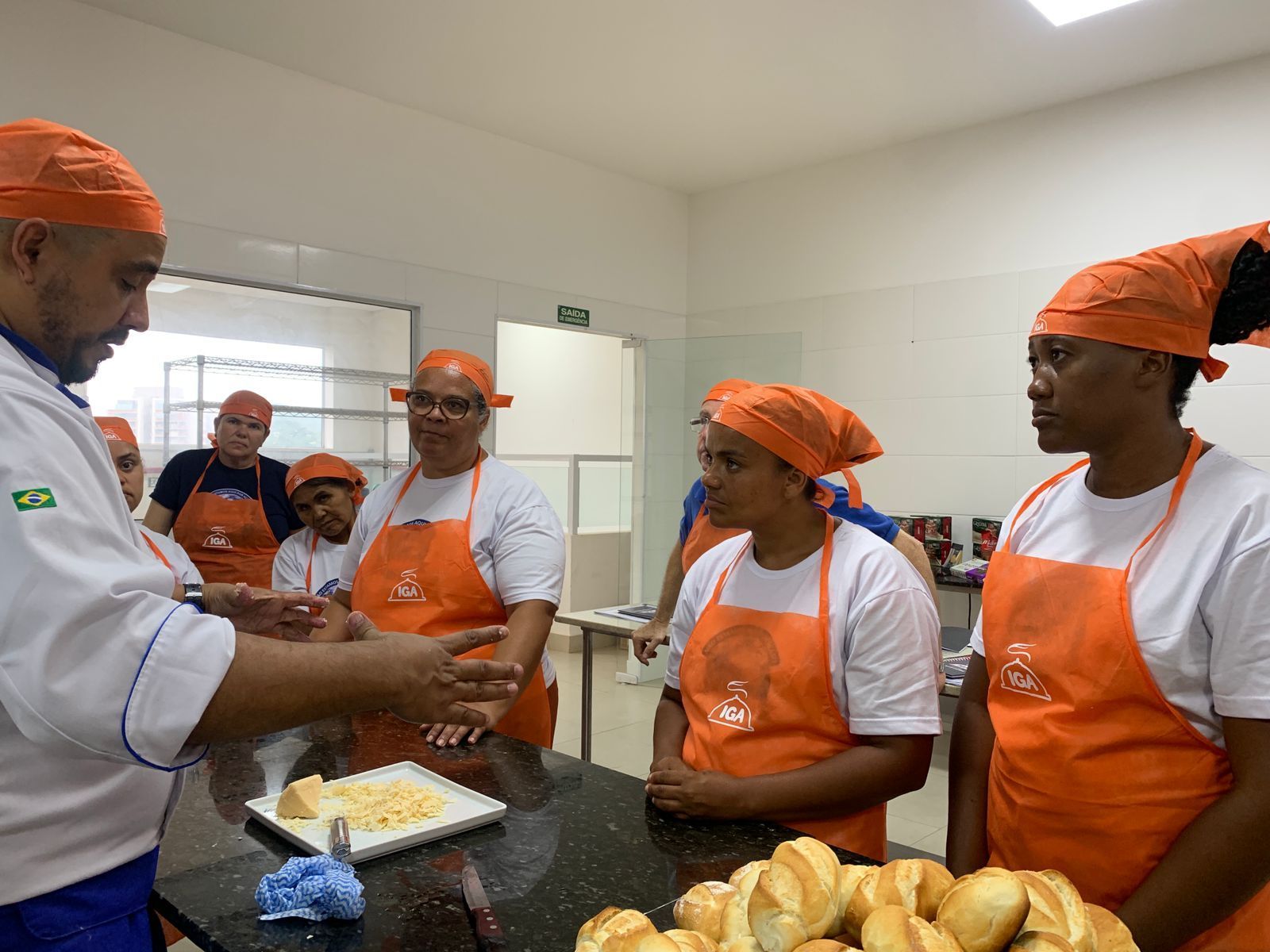Um chef instrui um grupo de pessoas vestindo aventais e lenços laranja na cabeça em uma cozinha, apontando para o queijo.
