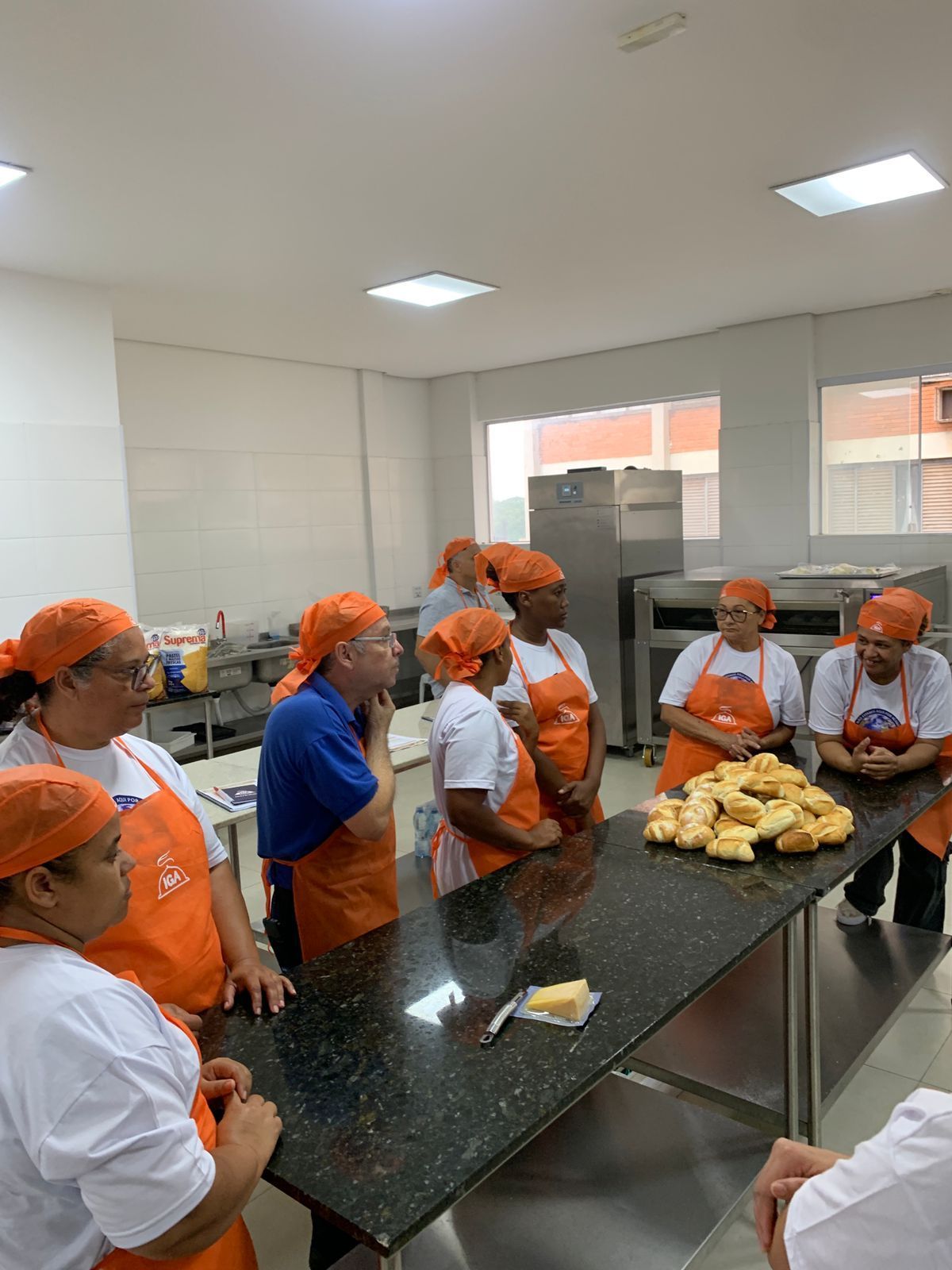 Um grupo de pessoas usando aventais e lenços na cabeça se reúne em volta de uma mesa com comida, em uma cozinha.