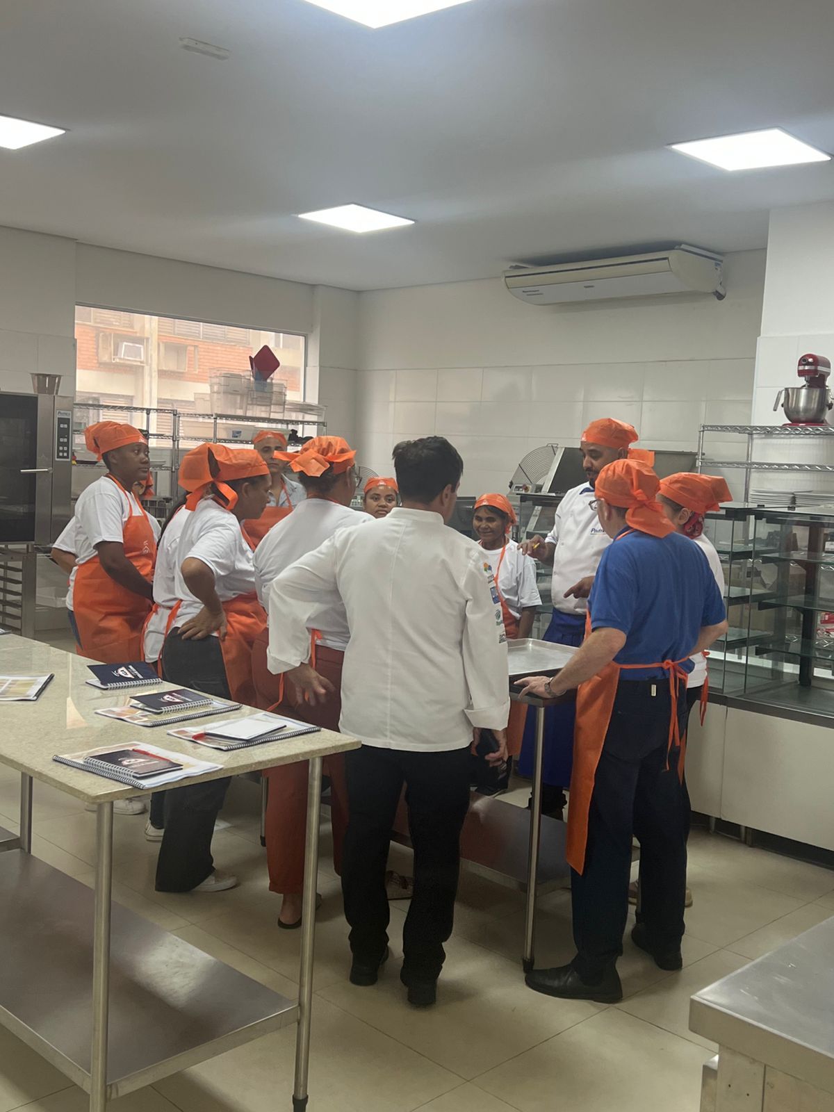Em uma cozinha industrial, pessoas, algumas usando aventais e bonés laranja, se reúnem em volta de uma mesa.