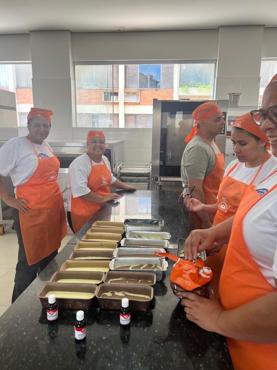 Pessoas com aventais e lenços na cabeça na cor laranja preparando alimentos em uma cozinha comercial, incluindo produtos de panificação.
