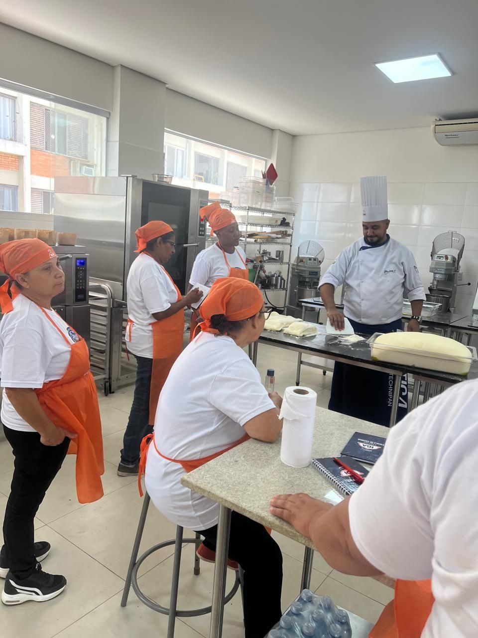 Chef ensinando técnicas de confeitaria a um grupo de pessoas usando aventais e lenços na cabeça em uma cozinha comercial.