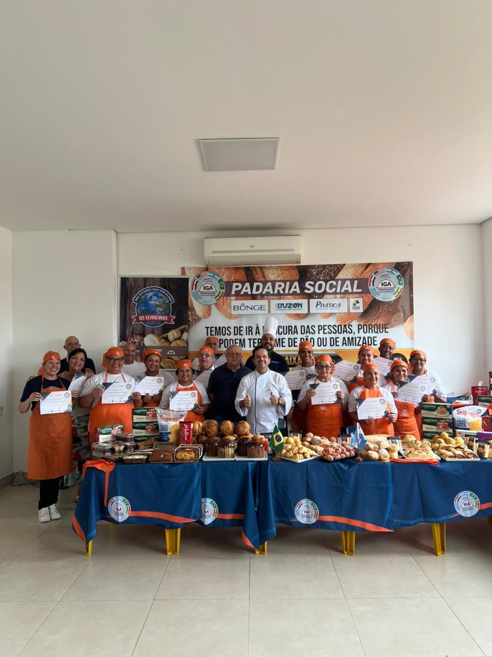 Um grupo de pessoas usando aventais e chapéus de chef está atrás de uma mesa repleta de comida, em frente a uma faixa.