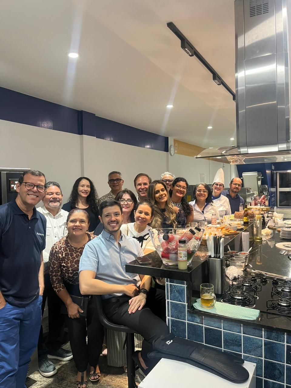 Grupo de pessoas sorrindo em frente a uma bancada de cozinha. Algumas pessoas seguram bebidas, e há comida visível.