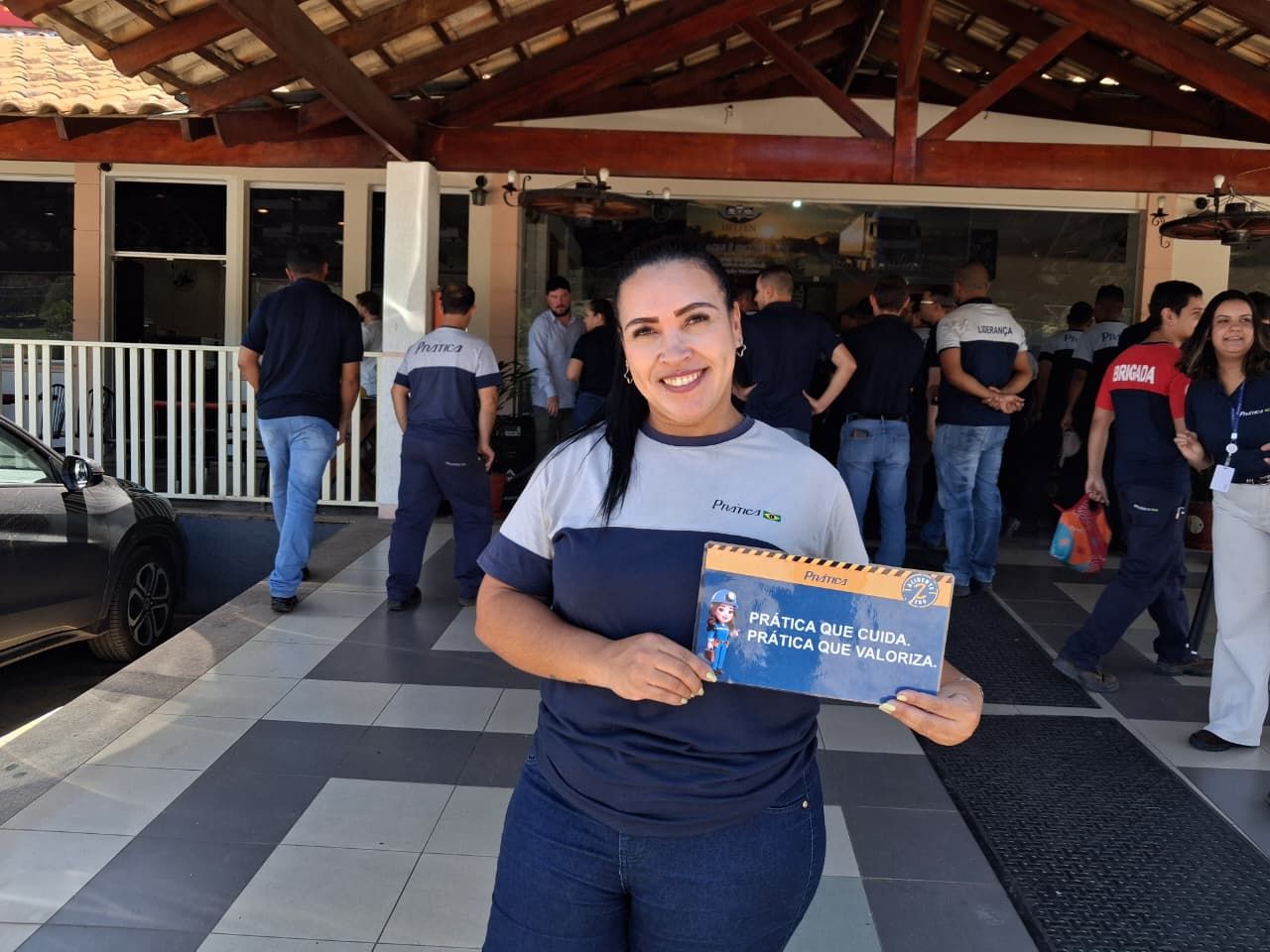 Uma mulher segurando uma placa sorri em frente a um prédio com pessoas. Ela veste um uniforme azul-marinho e cinza.