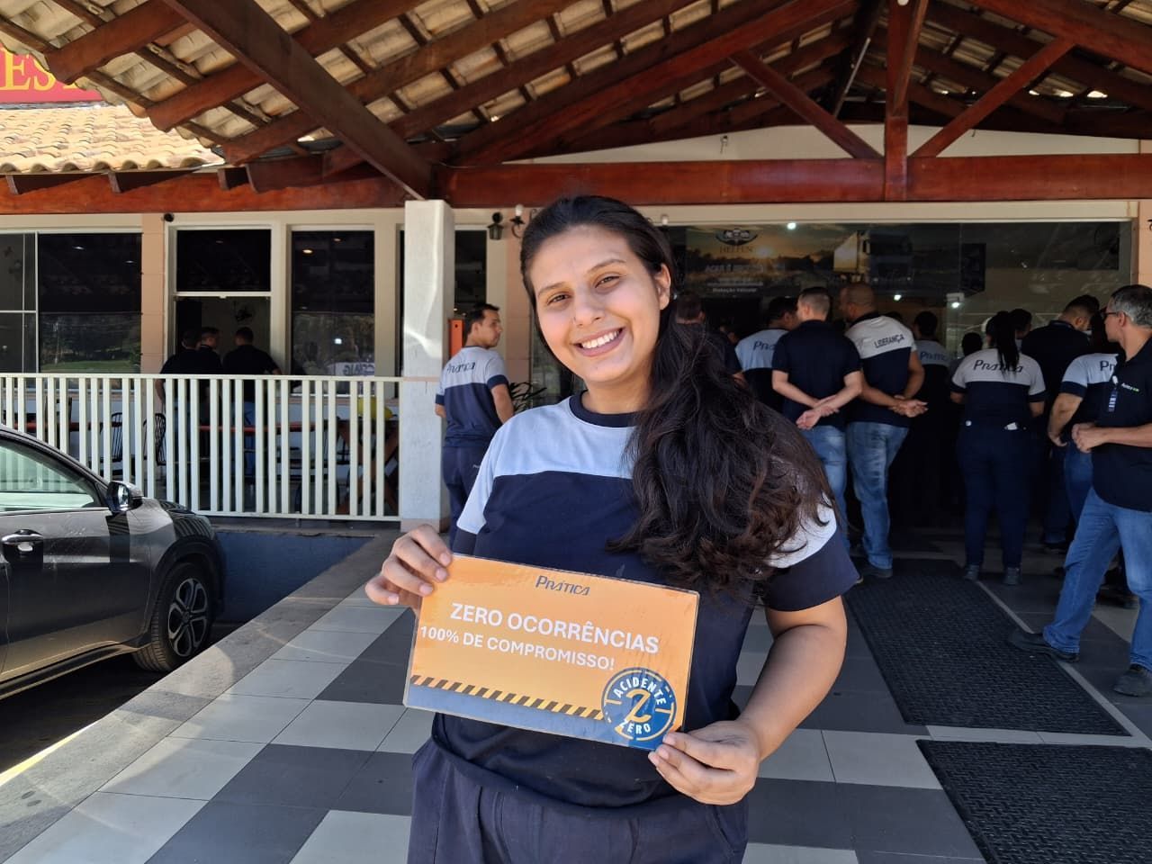 Mulher sorrindo, segurando uma placa do lado de fora de um prédio. Pessoas uniformizadas estão atrás dela.