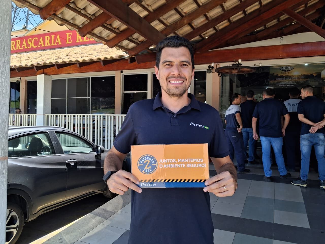 Homem sorrindo enquanto segura uma placa em frente a um restaurante. A placa é laranja e azul. Há pessoas ao fundo.