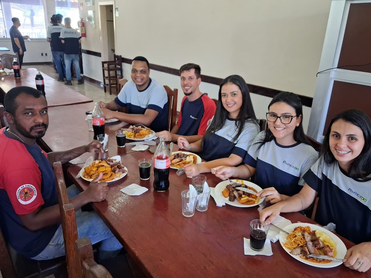 Pessoas comendo à mesa, algumas vestindo camisas azuis e brancas. Pratos de comida e bebida presentes.